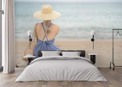 Woman in  blue dress and straw hat, sitting on a timber by the ocean. Wall mural