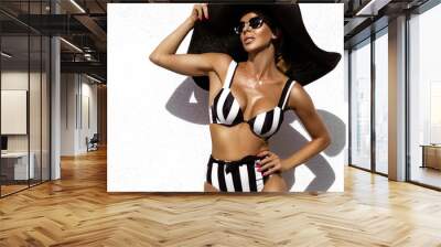 Elegant woman is wearing black and white striped bikini and big hat and is posing on white wall on summer day Wall mural