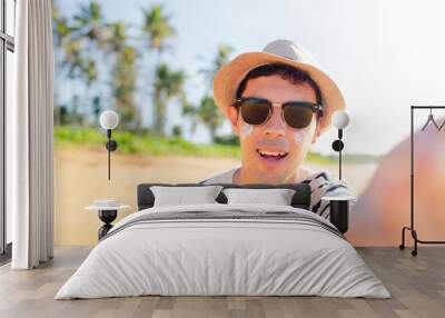 A man in beach wearing hat and sunglasses smiles, taking a selfie on a paradisiacal beach, capturing the essence of summer vacation joy. He is using sunscreen on his face. Wall mural