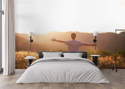 Young man sitting on the top of the mountain with his arms raised celebrating or thanking, with the sunset in the background. Concept of mental well-being, happiness, success and freedom. Wall mural