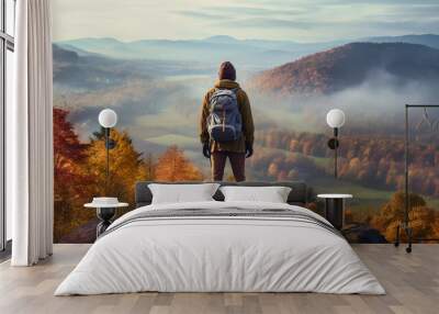 a hiker with a backpack standing on a cliff, overlooking a valley filled with autumn foliage, mist rising from the trees Wall mural