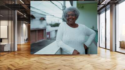 One happy elderly black woman from South America standing at humble residence balcony overlooking urban street in background. Portrait of 80s senior person with gray hair of African descent Wall mural