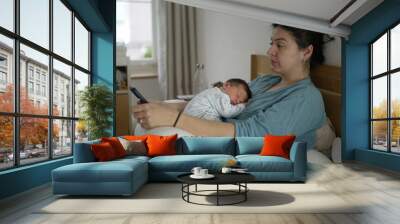 Tired mother with newborn baby sleeping on her chest, rubbing her eyes while holding a smartphone, wearing comfortable clothing, intimate indoor setting, showing exhaustion and love Wall mural