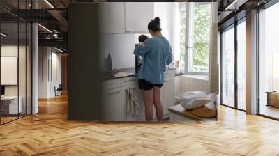 Woman in blue shirt holding baby in kitchen, multitasking between parenting and daily chores, illustrating the balance of motherhood and household responsibilities in a modern home Wall mural