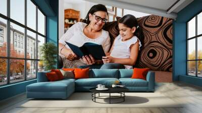 latin grandmother with granddaughter sitting on couch at home and reading book in Mexico city Wall mural