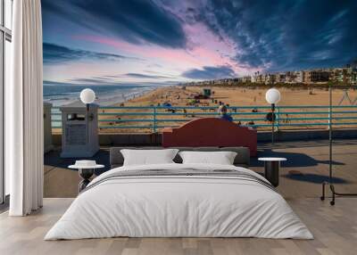 a man sitting on a red stone bench on the edge of the Manhattan Beach Pier with blue ocean water and people relaxing in the silky brown sand with homes and palm trees and powerful clouds at sunset Wall mural