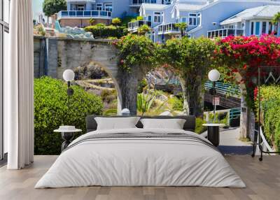 a shot of 3 stone arches with lush green plants and red flowers down a staircase with a white hand rail with gray homes along the mountain and blue sky at Dana Point Bluff Top Trail in Dana Point CA Wall mural