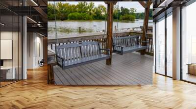 two gray wooden swings hanging from chains on a brown wooden deck over the Catawba River surrounded by rippling water and lush green trees at Kevin Loftin Riverfront Park in Charlotte North Carolina Wall mural