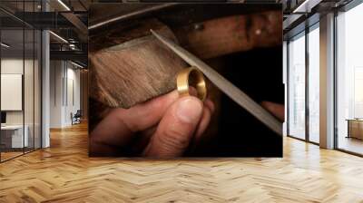 goldsmith hand holds a golden ring on the wooden workbench and works on it with a metal file, close up with copy space Wall mural