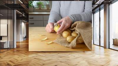 Hands of a woman are peeling raw potatoes on a wooden table in the kitchen at home Wall mural