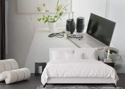 White office desk with laptop, camera, lenses, cable, glasses, notepad and a coffee cup, modern workplace of a photographer against a bright wall, copy space, selected focus Wall mural