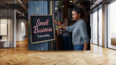 Smiling African American woman holding a 'Small Business Saturday' sign outside a shop. Concept for promoting local shopping and supporting small businesses during holiday events Wall mural