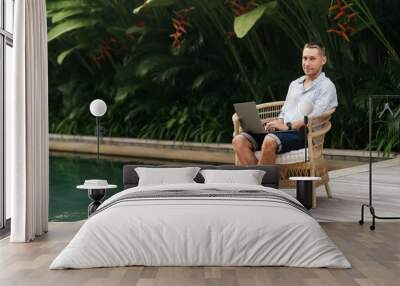 Young man using a laptop computer in a garden with a swimming pool. business, study, freelance Wall mural