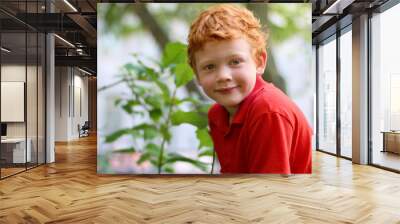 European boy with green eyes looking directly at the camera, close-up. Funny little child with curly ginger hair and freckles sitting on a tree. Fashion kid. Red casual shirt. Summer weather Wall mural