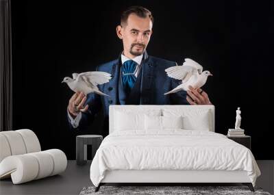 A magician in a blue tailcoat holds two white doves in his hands on a black background Wall mural