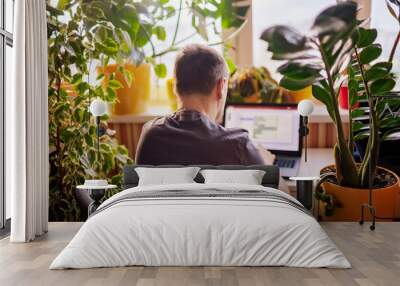 A man working with laptop remotely from home. A distant work place with many home plants. Green nature inspired home office. Indoor lifestyle Wall mural