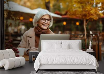 Aged woman uses her laptop in autumn park. Digital era, tech-savvy senior and modern technology Wall mural