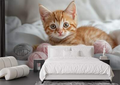 Adorable ginger kitten lounging on soft white bedding, surrounded by pastel pink and gray yarn balls, gazing curiously at the camera with wide eyes. Wall mural