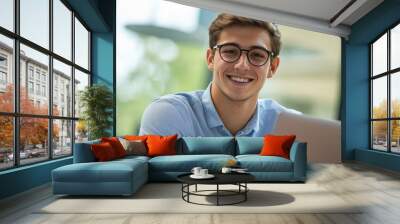 Cheerful young professional in light blue shirt and glasses smiles warmly at camera while working on laptop, embodying positivity and productivity in modern office setting. Wall mural