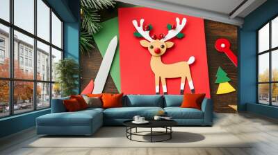 Festive DIY Christmas card with cute paper reindeer decoration, surrounded by craft supplies and holiday ornaments on rustic wooden background. Wall mural