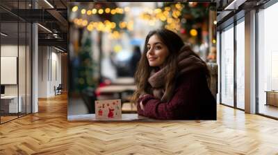 Festive winter scene: young woman in cozy sweater and scarf enjoys hot drink at outdoor cafe, surrounded by twinkling lights and holiday decorations. Wall mural