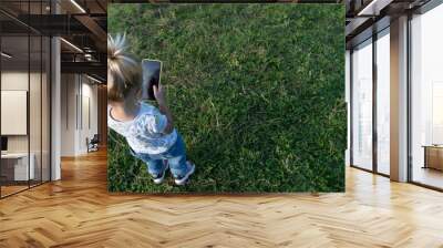 Little cute caucasian kid staying at green grass at summer, hanging mobile phone and playing online games or watching cartoons, modern digital generation of children, back view, copy space at grass. Wall mural