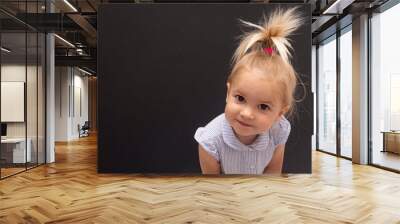 Portrait of cute little blond caucasian baby girl bending over in front of camera, smiling and looking the camera, black backgroung, copy space. Happy childhood concept Wall mural