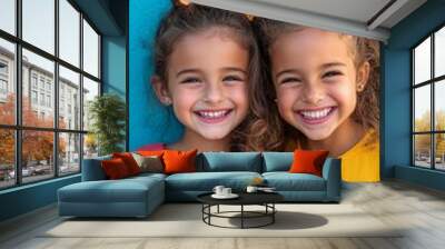 Two cheerful young girls with curly hair and bright smiles pose together against a vibrant turquoise wall, radiating joy and friendship in their colorful outfits. Wall mural