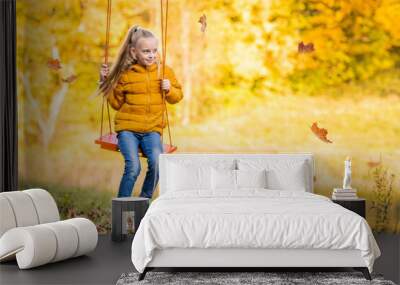 Happy little blonde caucasian girl smiling and riding a rope swing in autumn in the park. Wall mural