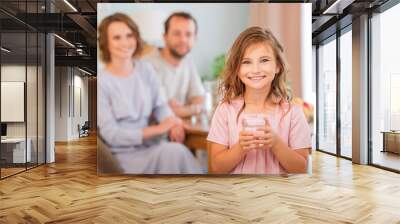 health and beauty concept - smiling little girl holding a glass of water. Happy parents in background. Wall mural