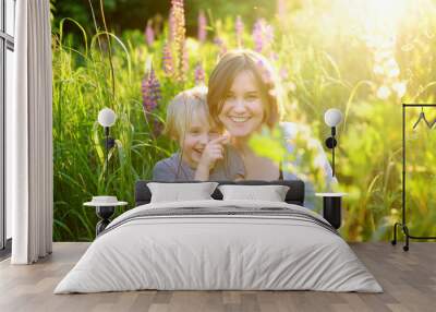 Charming woman with her little son walking in field of lupines on sunset. Outdoor activities for family. Wall mural