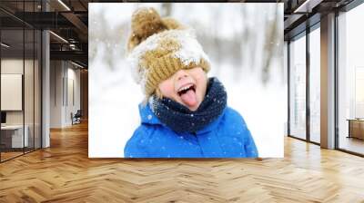 Funny little boy in blue winter clothes walks during a snowfall. Outdoors winter activities for kids. Wall mural