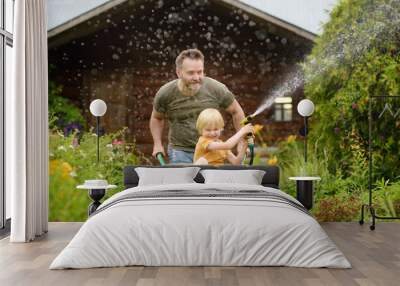 Happy little boy having fun in a wheelbarrow pushing by dad in domestic garden on warm sunny day. Child watering plants from a hose. Active outdoors games for kids in summer. Wall mural