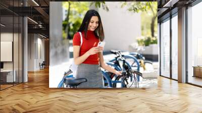 Beautiful smiling Indian woman holding mobile phone, reading text message  standing near bicycle on the street. Portrait of happy asian tourist in park. Travel, bicycle rental, summer concept Wall mural