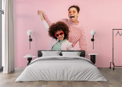 Charming mom and her young daughter in sunglasses and playfully posing isolated on pink background Wall mural