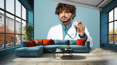 Handsome serious young Indian doctor holding stethoscope wearing white lab coat isolated on blue background in hospital. Portrait of medical student looking at camera. Medicine, health care concept Wall mural