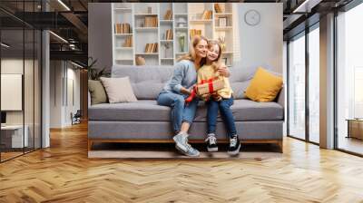 Mother's Day celebration at home: young daughter presents gift to mother, both sharing warm embrace Wall mural