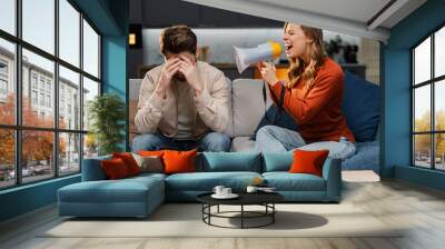 Portrait of angry young woman using, holding megaphone , screaming to man, shouting loudly while sitting on sofa in room at home. Concept of relationship, quarrel, conflict Wall mural