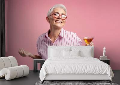 Portrait of mature gray haired female holding glass of alcohol looking at camera isolated on pink background. Smiling senior woman wearing stylish pink shirt, celebration success. Party time Wall mural