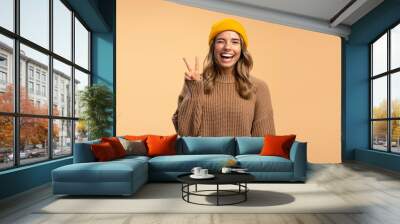 Portrait of smiling beautiful woman wearing stylish yellow hat and winter brown sweater showing victory sign looking at camera isolated on beige background, copy space. Concept of advertisement Wall mural