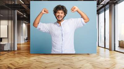 Smiling Indian man celebrating success with arms raised on blue background Wall mural