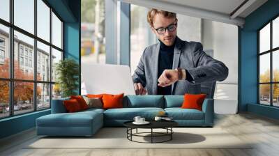 Young attractive businessman wearing stylish suit and eyeglasses  looking at watch check time working online in modern office   Wall mural