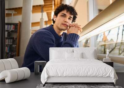 Young male student in earphones, listens to online lecture, sitting at desk with laptop in library Wall mural