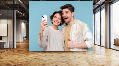 Young smiling friends hugging holding mobile phone making selfie over blue wall background Wall mural