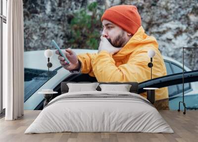 Young bearded man using phone while resting stop on road trip. He is wearing casual yellow jacket and red hat. Travel by car. Wall mural