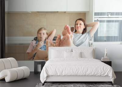 Cute girl and her mother are smiling while eating ice cream in the kitchen with legs on a table Wall mural