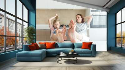 Cute girl and her mother are smiling while eating ice cream in the kitchen with legs on a table Wall mural