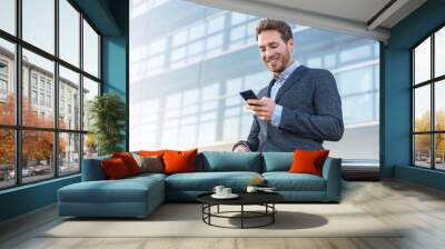 Businessman using mobile phone app texting outside of office in urban city with skyscrapers buildings in the background. Young caucasian man holding smartphone for business work. Wall mural