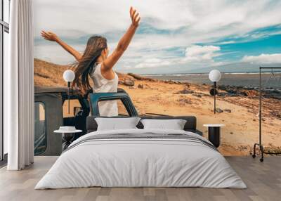 Car road trip travel fun happy woman tourist with open arms at ocean view from sports utility car driving on beach. Summer vacation adventure girl from the back. Wall mural