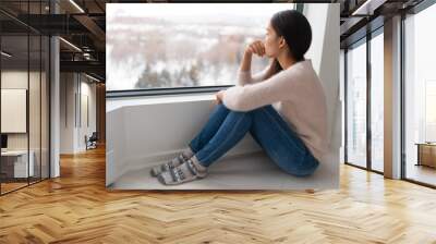 Depressed young girl feeling sad an lonely, anxious looking out the window in winter. Unhappy Asian woman alone, depression at home. Wall mural